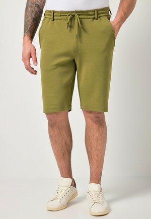 Mand iført olivengrønne shorts med snøre og hvide sneakers, stående med hænderne tæt på lommerne, synlig tatovering på venstre arm.