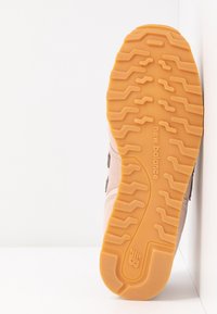 Semelle en gomme d'une chaussure New Balance montrant le motif de la semelle et le logo, positionnée à la verticale contre un fond blanc en angle.