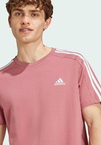 T-shirt rosa Adidas con design a tre strisce bianche sulle spalle, scollatura rotonda e un piccolo logo sulla parte sinistra del petto.