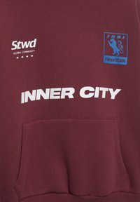 Sweat à capuche bordeaux avec une poche avant ; présente un texte blanc "INNER CITY" et un graphique bleu avec "1991" et un lion, accompagné de "Stwd GLOBAL COMMUNITY."