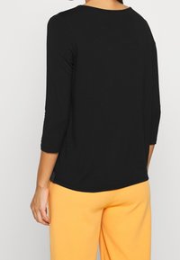 Svart, långärmad topp med rundad halsringning och mjuk textur, med en avslappnad passform och sidslitsar, kombinerad med orangea byxor.