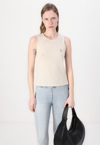 Beige ermeløs tank topp laget av lett stoff, med en subtil logo på brystet, paret med lyseblå skinny jeans og en svart skulderveske.