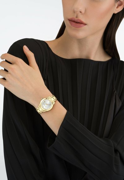 Mujer que lleva una blusa negra de manga larga plisada y un reloj de pulsera dorado marcando las 10:10 en la muñeca izquierda, con la mano apoyada en el hombro derecho.