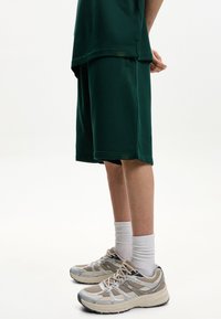 Zelene bombažne kratke hlače z ohlapnim krojem, v kombinaciji s svetlobezhnimi športnimi copati in belimi nogavicami do gležnjev, prikazujejo gladko teksturo in preprost dizajn.