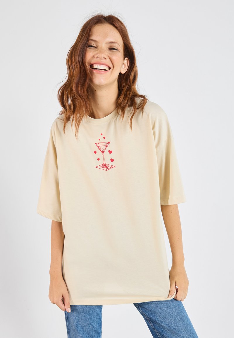 Beige oversized tričko s červenou grafikou martini sklenice srdíček. Vyrobeno z měkkého materiálu, s krátkými rukávy a kulatým výstřihem.