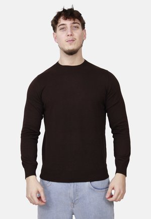 Giovane uomo con maglione nero aderente a maniche lunghe e jeans azzurri, in piedi davanti a uno sfondo bianco uniforme.