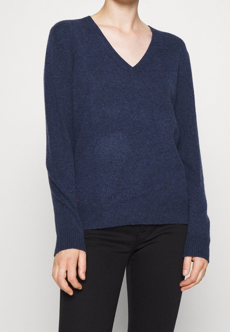 Personne portant un pull à manches longues bleu marine à col en V associé à un pantalon noir, sur un fond clair uni.