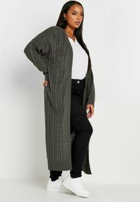 Lång, mörkgrön stickad cardigan med kabelstickning, öppen framside-design, ribbad fåll och ärmslut, i kombination med en vit V-ringad t-shirt och svarta byxor.