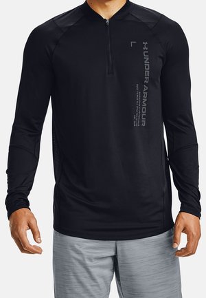 Mężczyzna ubrany w czarną długą koszulkę sportową Under Armour z zamkiem błyskawicznym i szare spodenki, stojący na jednolitym tle.