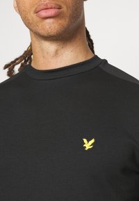 Felpa nera girocollo con una superficie liscia e un piccolo logo di un uccellino giallo sul lato sinistro del petto. Presenta accenti tonali sulle spalle.
