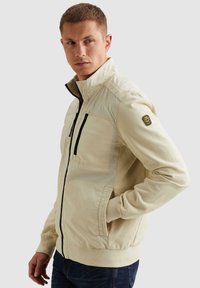 Homme portant une veste beige zippée avec des accents noirs sur la fermeture éclair et un écusson sur la manche, debout la main dans la poche, regardant sur le côté.