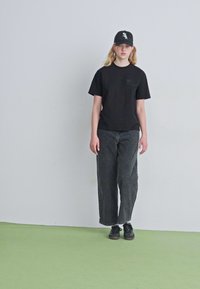 T-shirt de algodão preto, jeans de corte solto cinza escuro, ténis pretos e um boné preto com um logótipo, em frente a uma parede clara e um chão verde.