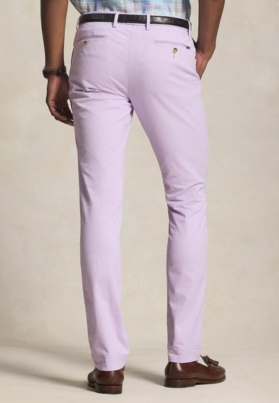 Άνδρας που φοράει ανοιχτό μωβ στενά παντελόνια, καφέ δερμάτινη ζώνη και καφέ μοκασίνια με φούντες, στέκεται μπροστά σε ουδέτερο φόντο.