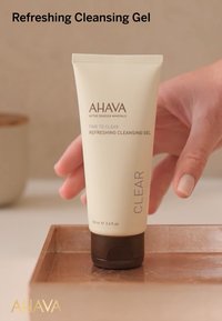 Gel limpiador refrescante AHAVA en un tubo beige con una tapa marrón, mostrando el nombre de la marca y los detalles del producto, sostenido sobre una bandeja de madera.