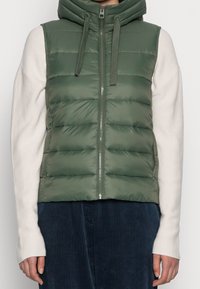 Gewatteerd vest in olijfgroen met capuchon, met horizontale doorgestikte lijnen, een ritssluiting aan de voorkant en geribde crèmekleurige mouwen, gecombineerd met donkergroene ribfluwelen broek.