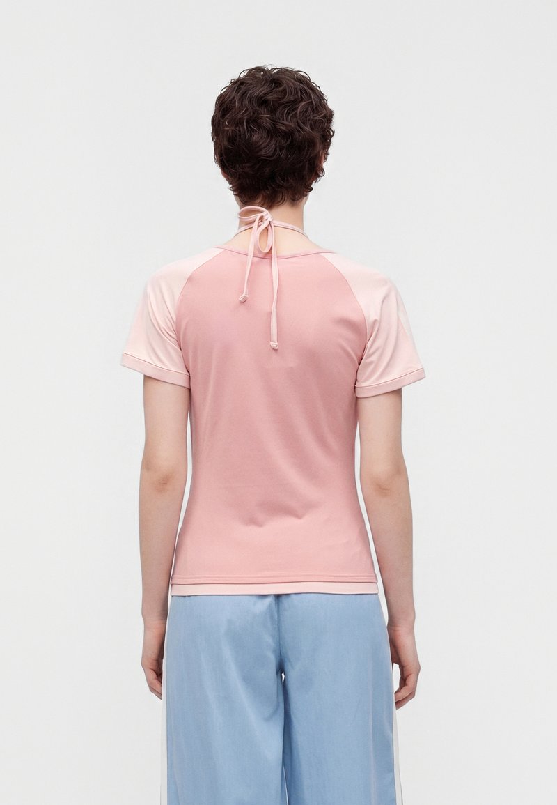 Personne aux cheveux courts et bouclés portant un haut rose à manches courtes avec liens au cou et un pantalon bleu clair, vue de dos.
