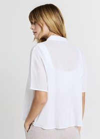 Blouse blanche à manches courtes en tissu texturé, avec une coupe décontractée et un ourlet arrondi. L'encolure arrière est légèrement plus haute, avec une légère brillance.