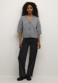 Grijze gebreide cardigan met korte mouwen, voorzien van decoratieve bloemenknopen, een V-hals en een geribbelde zoom. Gepaard met donkerdere rechtlopende jeans.