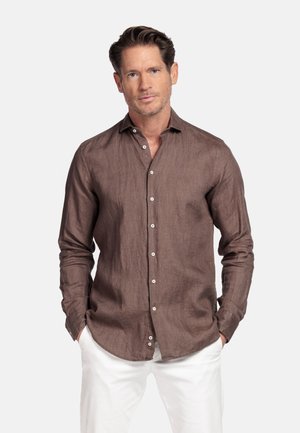 Man met een bruin overhemd en witte broek, staand met handen in de zakken tegen een effen achtergrond.