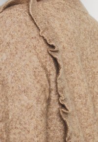 Tissu en mélange de laine de couleur brun clair avec une finition texturée. Présente un détail de bord froncé le long de l'encolure, ajoutant de la profondeur au design.