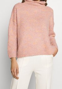 Chunky rosa tröja med en hög, vikbar krage, korta ärmar och ett ribbat avslut, som har inslag av orange fläckar genom hela stickningen.