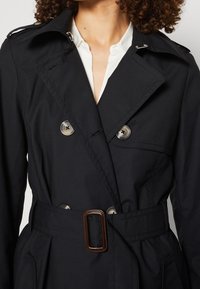 Svart trenchcoat i slätt tyg, med en dubbelknäppt design, stora metallknappar och ett bälte med en rektangulär spänne.