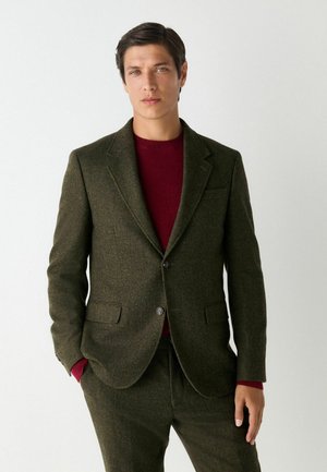 Blazer cu model herringbone de culoare verde închis, cu revere cu colț, două buzunare frontale și închidere cu un singur nasture. Purtați peste un pulover burgundy.