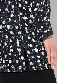 Blouse en tissu noir présentant un motif de cercles blancs et beiges, avec une fente latérale et des manches courtes, mettant en valeur une surface douce et texturée.