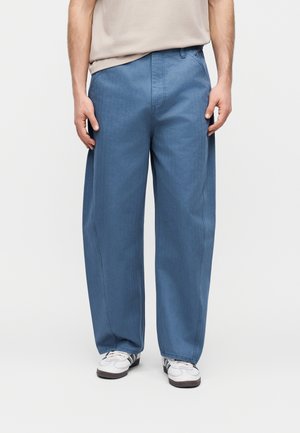 Mężczyzna w luźnych niebieskich spodniach i białych tenisówkach stoi na tle jednolitego, jasnego tła.