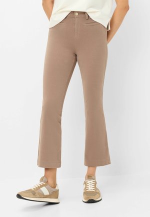 Person trägt beige gekürzte Schlaghosen mit aufgesetzten Fronttaschen und beige-weiße Schnürsneaker vor weißem Hintergrund.