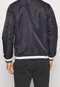 Černá bomber bunda s hladkým, lesklým povrchem. Obsahuje bílý a černý pruhovaný ribbed lem a manžety. Bez viditelného značení.