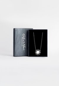 Sølv halskæde med cirkulært vedhæng, der har en glat, reflekterende overflade, omkranset af et geometrisk metaldesign, præsenteret i en mørk æske.