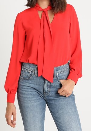Femme portant un chemisier rouge à manches longues avec un foulard noué autour du cou et un jean bleu taille haute, une main dans la poche.