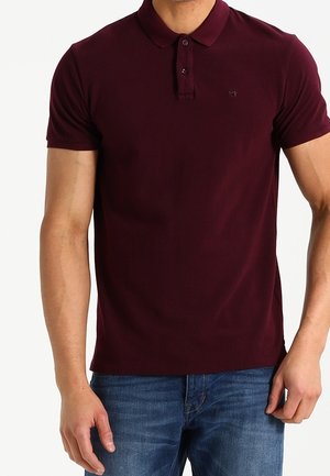 Homme portant un polo bordeaux ajusté à manches courtes et un jean bleu, debout devant un fond blanc uni.