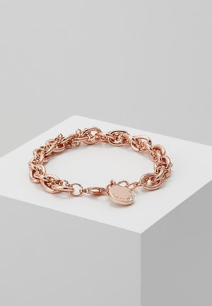 Bracelet chaîne en or rose avec des maillons ovales et un charm rond en forme de cœur sur le fermoir, présenté sur un socle blanc sur fond gris.