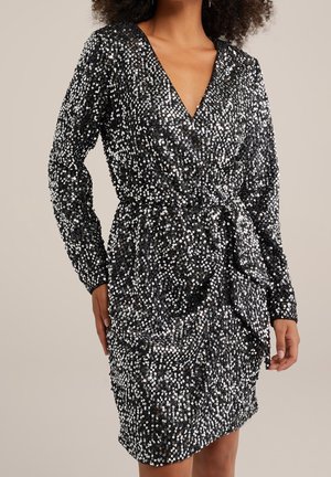 Vrouw draagt een lange mouwen, zwarte wikkeljurk die volledig bedekt is met reflecterende zilveren pailletten, staand met handen langs haar zij.