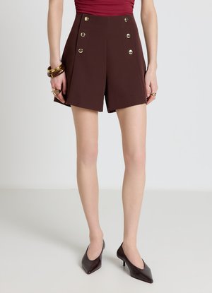 Donna che indossa pantaloncini marroni a vita alta con sei bottoni dorati, abbinati a tacchi marroni, braccialetti e anelli dorati su uno sfondo neutro.