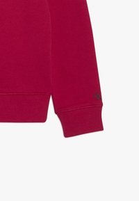 Roter Langarm-Pullover mit geripptem Saum und kleinem schwarzen "ck"-Logo nahe dem Handgelenk auf weißem Hintergrund.