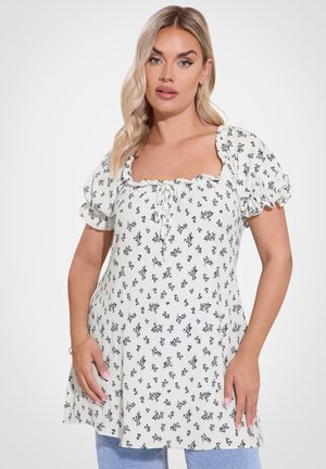 Blondynka ubrana w białą, kwiecistą bluzkę z krótkim rękawem o kwadratowym dekolcie i bufkami na rękawach, w połączeniu z jasnoniebieskimi dżinsami.