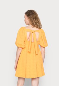 Vestido amarelo, texturizado, com mangas bufantes e costas abertas atadas com um laço. Forma em A que chega até à metade da coxa. Sem padrões ou acentos visíveis.