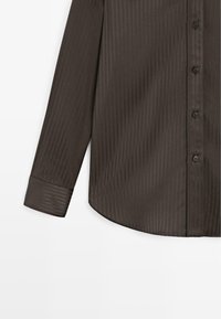 Camicia a maniche lunghe in tessuto marrone scuro con sottili righe verticali, dotata di chiusura anteriore con bottoni e orlo curvo.