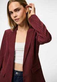 Woman with straight blonde hair wears a maroon pinstripe blazer, white cropped top, and blue jeans, touching her ear against a white background.