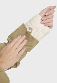 Veste kaki avec une doublure intérieure crème, dotée d'une fermeture à pression au niveau des poignets et d'une texture douce et matelassée.