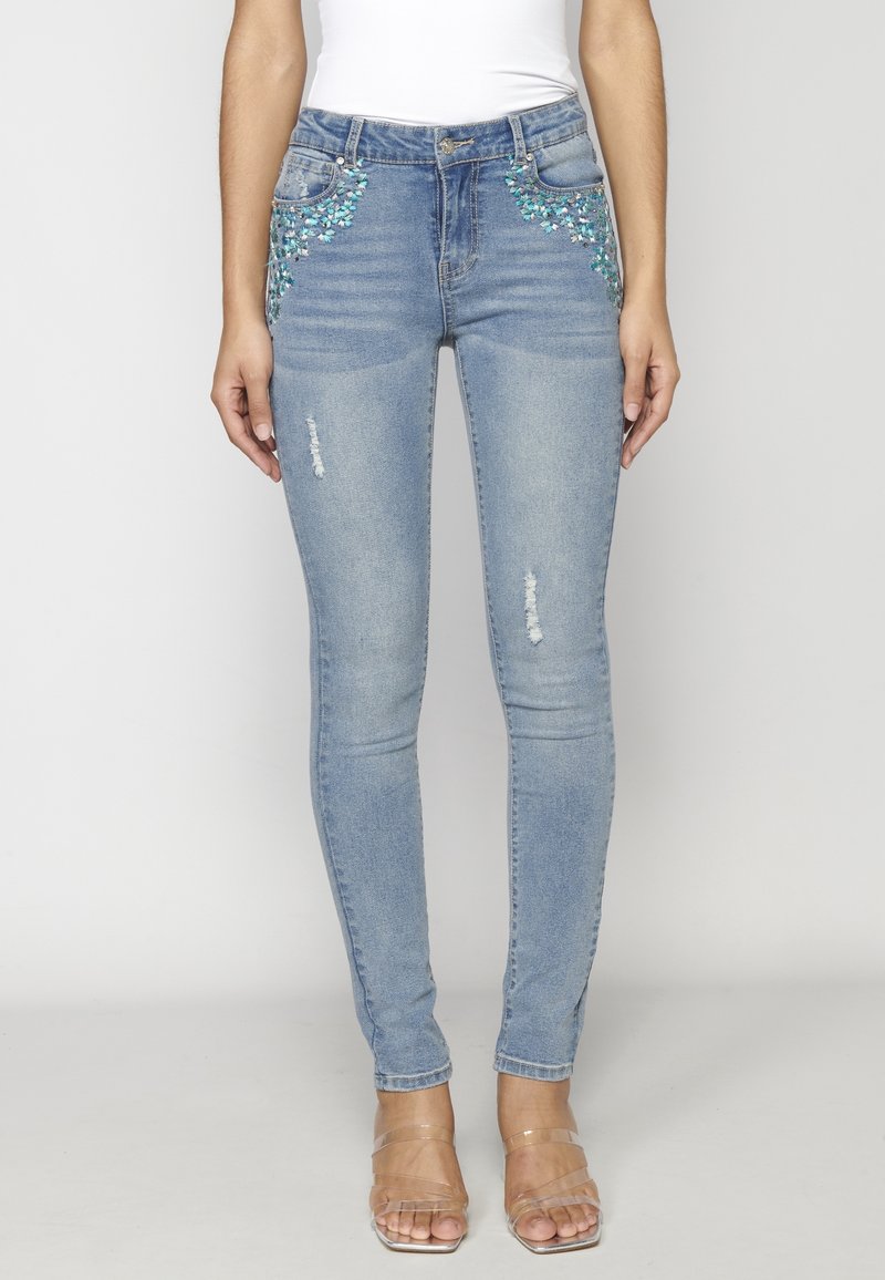 Jeans ajustados de color azul claro con parches deslavados y adornos decorativos en turquesa en los bolsillos. Detalles desgastados cerca de la zona de las rodillas.