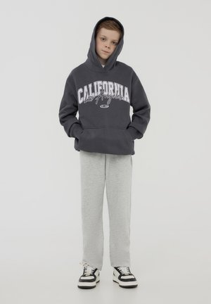 Felpa grigia con stampa "California Los Angeles", abbinata a pantaloni della tuta grigi chiari. Sneakers in bianco e nero completano l'outfit.