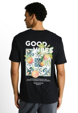 Mann von hinten, trägt ein schwarzes Shirt mit dem Schriftzug "GOOD VIBES" und bunten Pfirsichen über einem dekorativen Fliesenmuster auf dem Rücken.