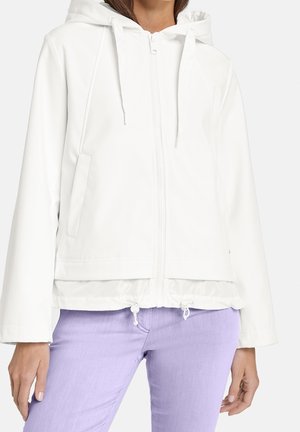 Femme portant une veste blanche zippée à capuche avec cordons et un pantalon violet clair, debout sur un fond uni.