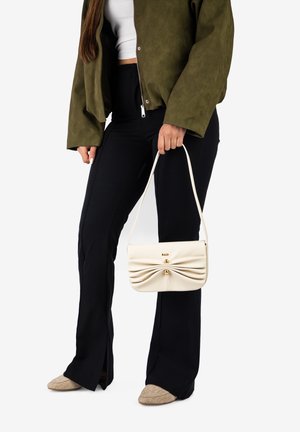 Woman wearing olive jacket, white crop top, black pants, beige shoes, holding cream handbag with gathered design and gold accents.