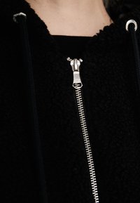 Černá fleecová bunda se zipem, která má stříbrný zip a kapuci na šňůrku. Textura vypadá měkká a huňatá.