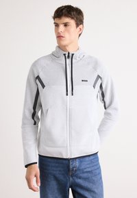 Veste zippée gris clair en tissu texturé, avec des accents noirs et un col haut. Présente un logo sur la poitrine et un design de panneaux contrastants.